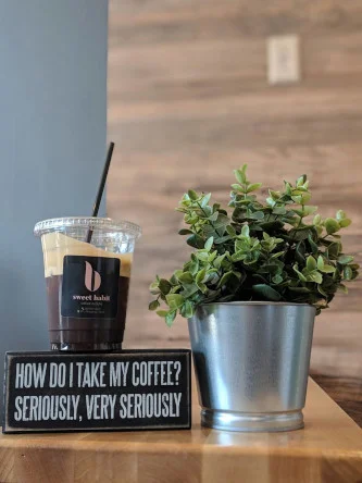 An iced coffee in a branded cup beside a potted plant and a sign asking how you take your coffee