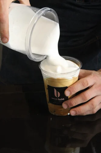 A barista pours steamed milk from a pitcher into an iced coffee in a Sweet Habit cup