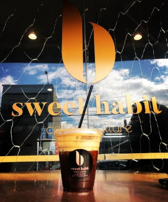 An iced coffee held up against the shop window with the Sweet Habit sign and orange b mark behind it