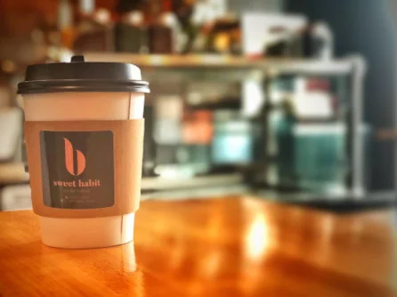 A coffee cup with a Sweet Habit sleeve on the wood counter