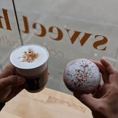 Two hands holding a dusted latte and a powdered doughnut in front of the shop window