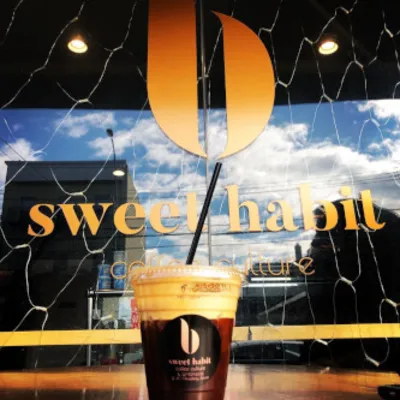An iced coffee held up against the shop window with the Sweet Habit sign and orange b mark behind it